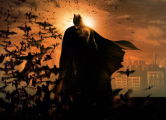 Batman begins character standing with bats flying around in a dark cityscape at sunset