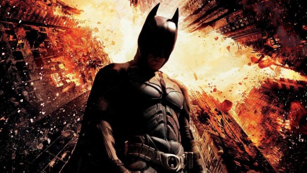 Batman begins character stands amid fiery destruction in the cityscape background