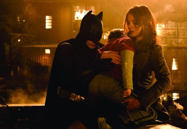Batman Begins holding a child while a woman looks on in a dimly lit industrial setting