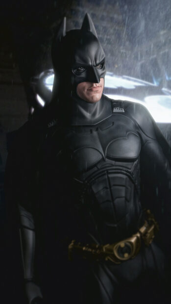 Batman Begins character standing in rain with bat symbol glowing behind