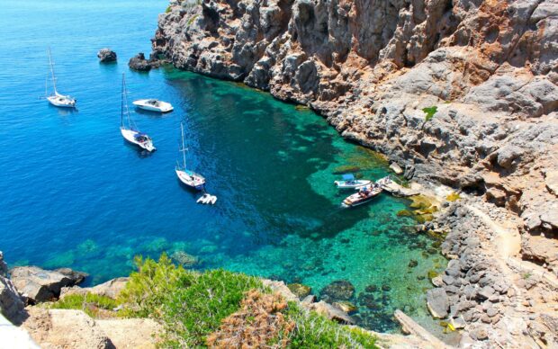 A clear blue sea with rocky cliffs and boats anchored near the shore in Spain, 2K Desktop Wallpaper