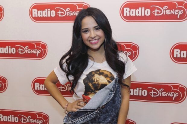 Becky G wearing denim overalls and a graphic t shirt smiling at a Radio Disney event