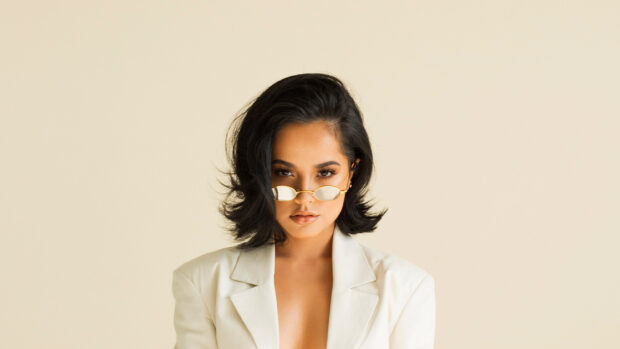 Becky G wearing small sunglasses and a white blazer looking confidently at the camera