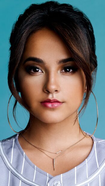 Becky G wearing large hoop earrings and layered necklaces with a striped top against a blue background