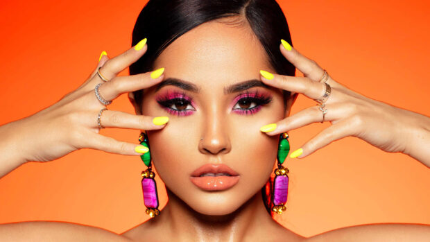 Becky G with colorful earrings and vibrant makeup posing against an orange background