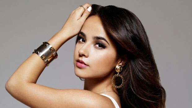 Becky G with long brown hair and gold jewelry posing with her hand on her head