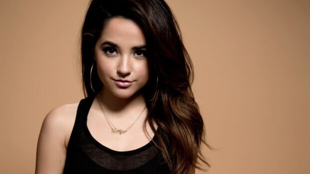 Becky G posing with long hair and hoop earrings in a black top against a beige background
