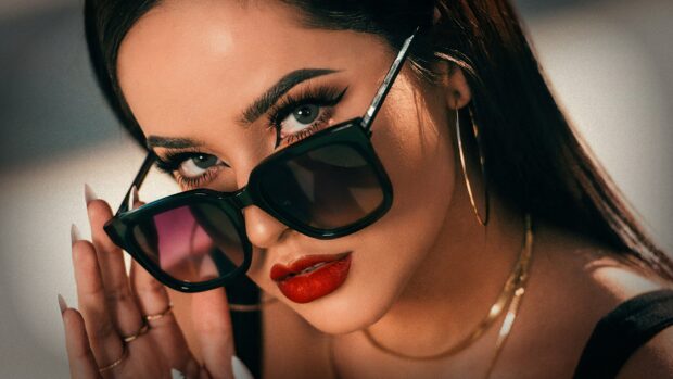 Becky G wearing large sunglasses and red lipstick in a close up portrait
