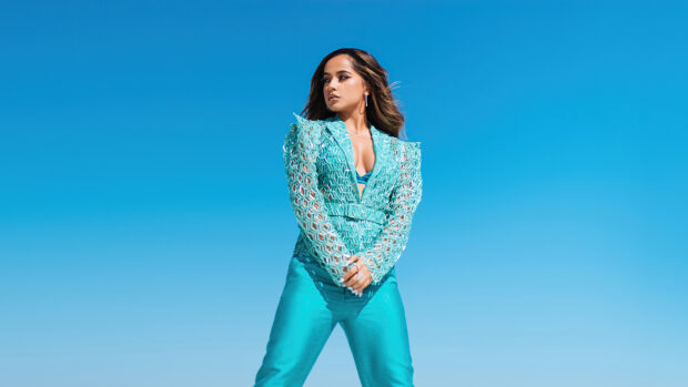 Becky G wearing a stylish turquoise outfit standing confidently against a clear blue sky