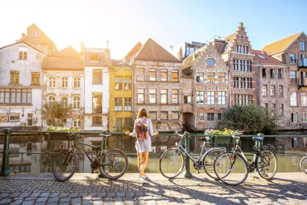 A woman with a backpack stands near bicycles by the canal in Belgium cityscape