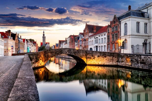 Historic Belgium architecture along a calm canal with a stone bridge at sunset