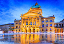 The illuminated Federal Palace building is reflected on wet ground in Bern Switzerland at dusk