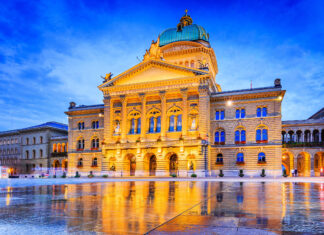 The illuminated Federal Palace building is reflected on wet ground in Bern Switzerland at dusk