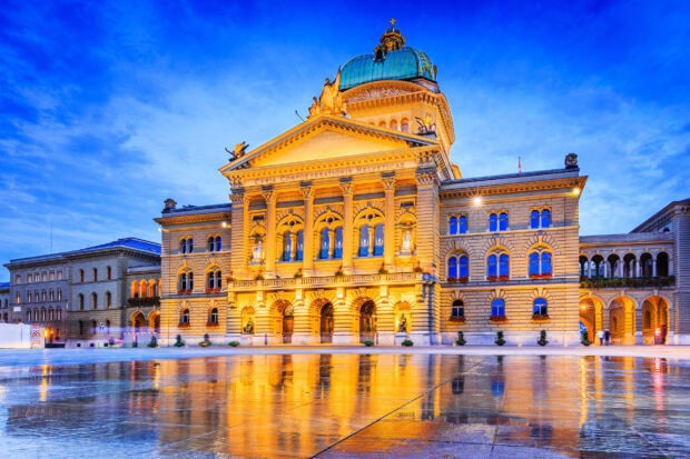 The illuminated Federal Palace building is reflected on wet ground in Bern Switzerland at dusk