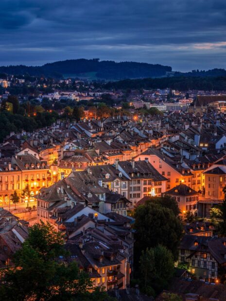 Bern cityscape at dusk with illuminated historic buildings and hills under a cloudy sky
