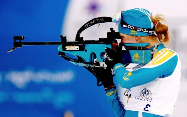A biathlon athlete aiming with a rifle during a competition wearing blue and white gear
