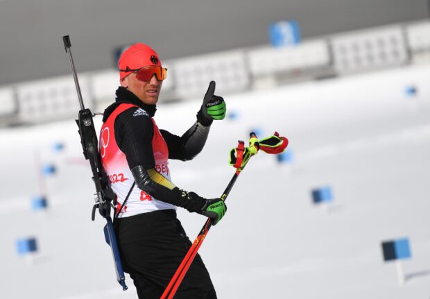 A biathlon athlete wearing red and black gear giving a thumbs up during a winter competition