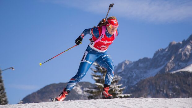 Female biathlon athlete skiing on snow with mountain background under clear sky