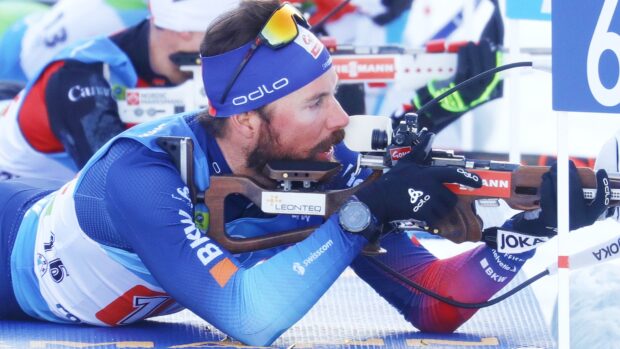 A biathlon athlete aiming with a rifle while lying down during a competition in snow