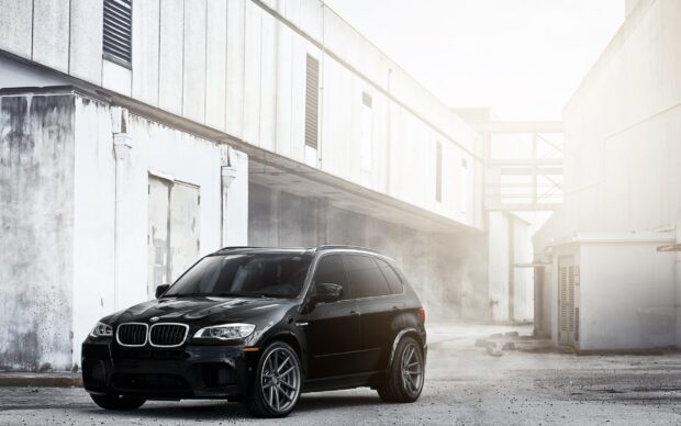 A black BMW X5 is parked in a bright industrial alley with white buildings and a misty atmosphere, Desktop Wallpaper
