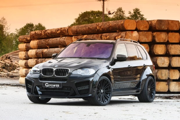 A black BMW X5 is parked in front of a large stack of cut wooden logs at sunset, 2K Desktop Wallpaper