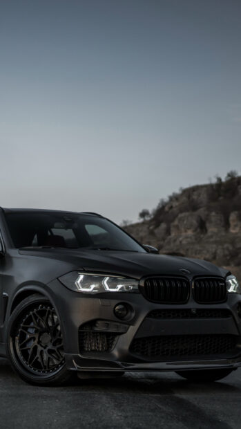 A sleek black BMW X5 with illuminated headlights is positioned on a road near rocky terrain, 4K Wallpaper