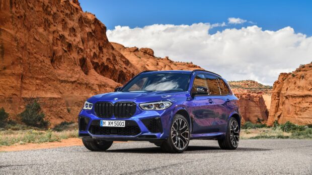 A blue BMW X5 is parked on a paved area with red rock cliffs and green shrubs in the background