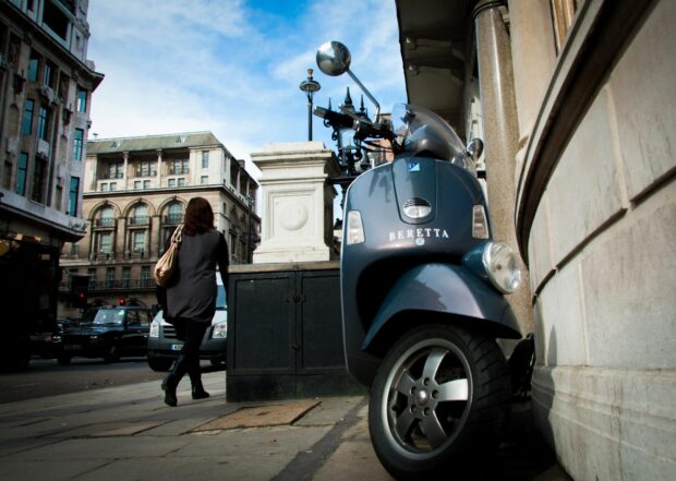 A blue scooter is parked against a building on a busy city street with a pedestrian walking by, HD Desktop Wallpaper