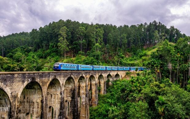 A blue train is traveling over a stone arch bridge surrounded by lush greenery in Sri Lanka, 2K Desktop Wallpaper