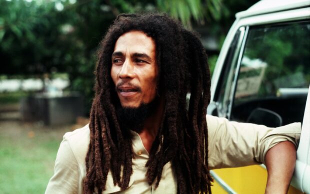 A man with dreadlocks is standing beside a yellow and white vehicle looking thoughtfully, 2K Desktop Wallpaper