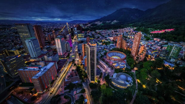 The Bogota cityscape at twilight shows illuminated buildings and traffic with mountains in the backgroung
