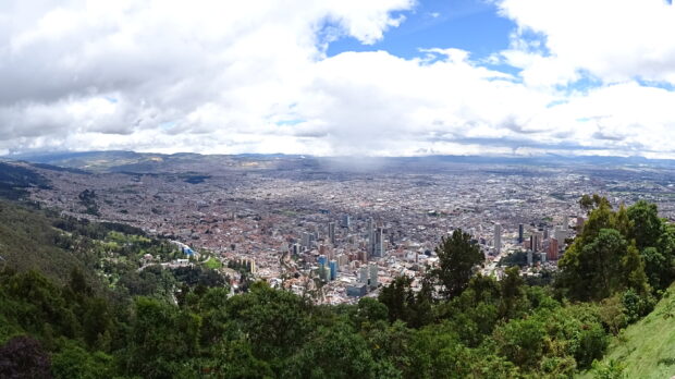 A panoramic view of Bogota shows the city surrounded by green trees and cloudy sky, 2K Desktop Wallpaper