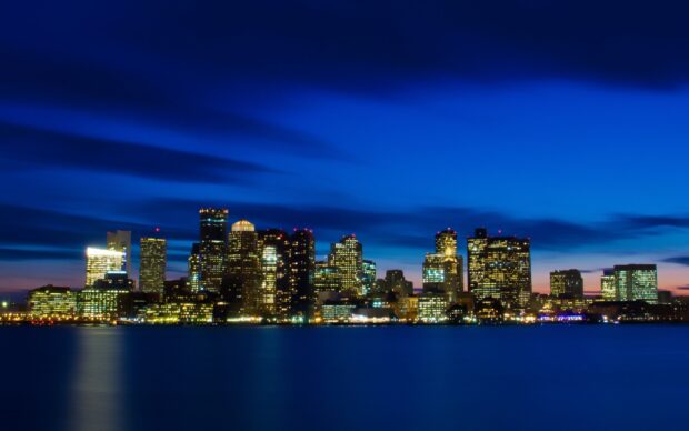 Boston skyline illuminated at night with clear blue sky and calm water in front