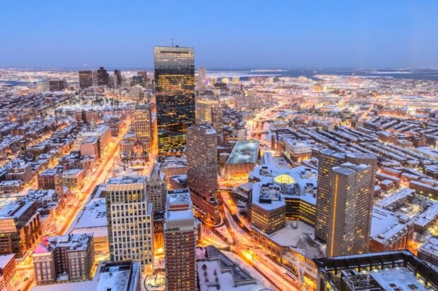A snowy cityscape of Boston with illuminated streets and tall buildings at dusk