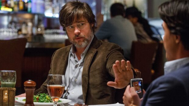 Brad Pitt sitting at a restaurant table with glasses and salad wearing a brown jacket