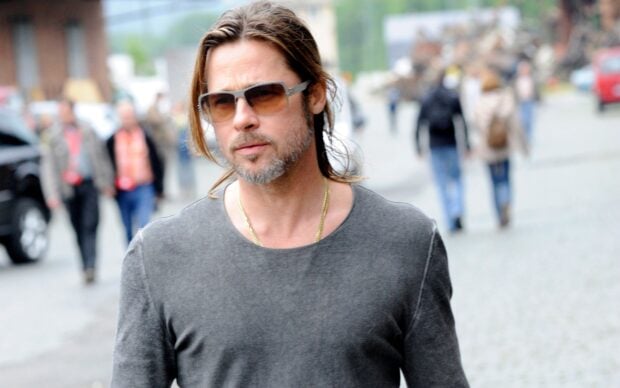 Brad Pitt wearing sunglasses and a gray shirt walking outdoors on a street with blurred people in the background