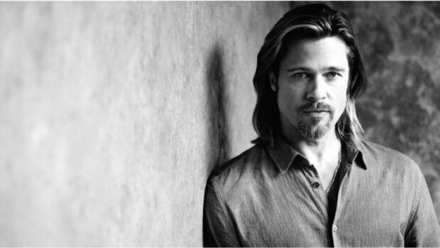 Brad Pitt standing against a textured wall with long hair and a goatee in black and white