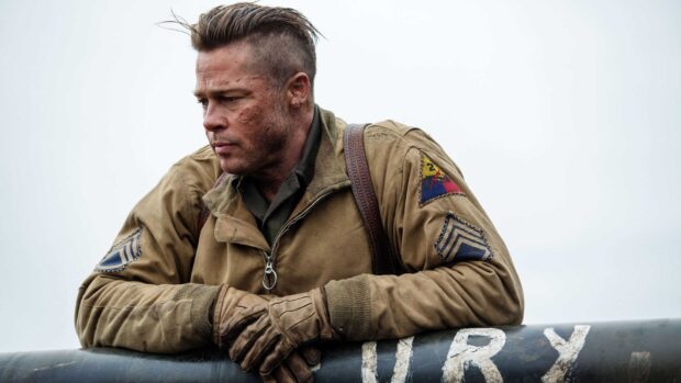 Brad Pitt wearing a military jacket and gloves leaning on a plane in a war setting