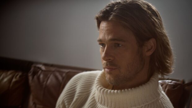 Brad Pitt wearing a white knitted sweater sitting thoughtfully on a brown leather couch
