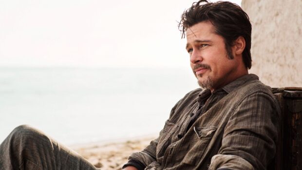 Brad Pitt sitting thoughtfully on the beach in a casual outfit