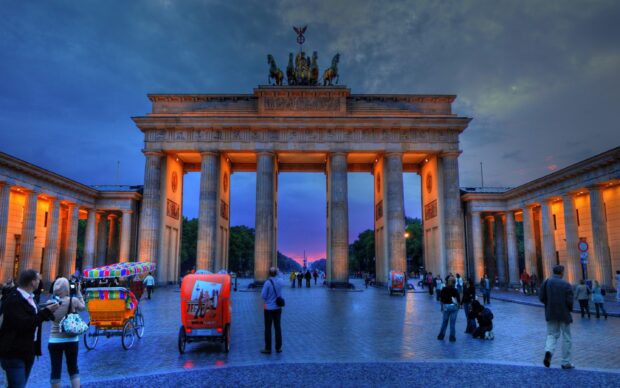 The Brandenburg Gate in Berlin stands illuminated with people and rickshaws nearby at dusk, 2K Desktop Wallpaper