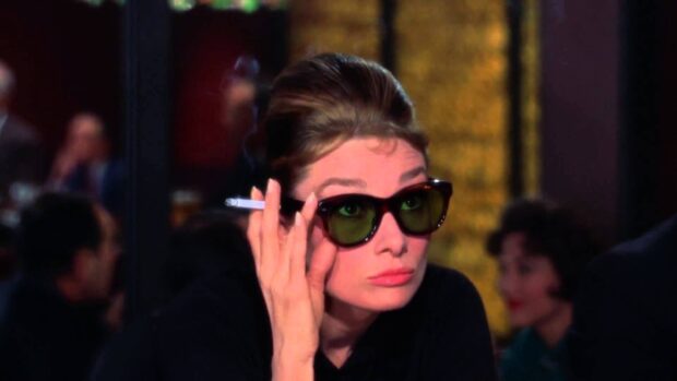 A woman from Breakfast At Tiffanys wearing dark sunglasses and holding a cigarette in a cafe