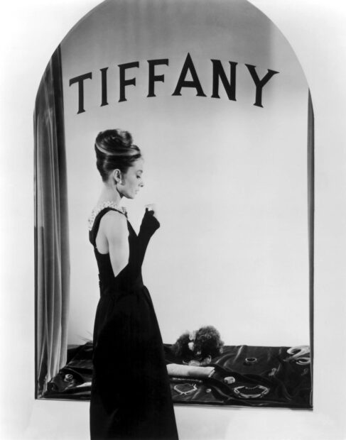 A woman wearing a black dress and gloves stands in front of a Tiffany display window