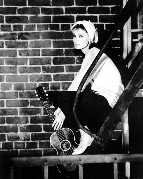 A woman wearing a headscarf holding a guitar sits on metal stairs against a brick wall