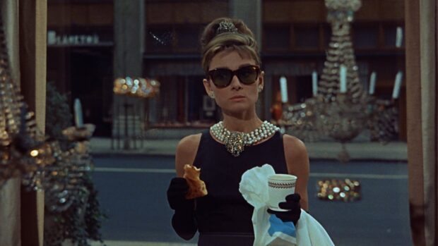 A woman wearing pearls and sunglasses eating pastry with a coffee cup in the scene from Breakfast At Tiffanys