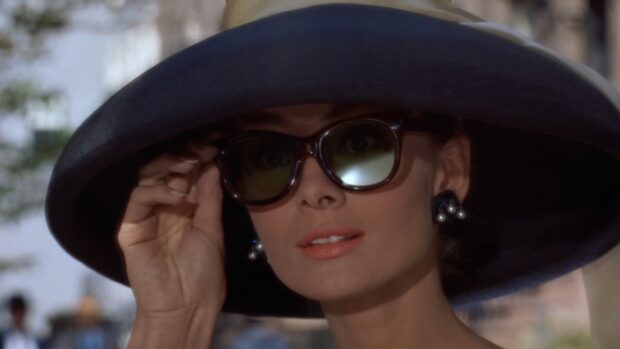 A woman wearing sunglasses and a large hat from Breakfast At Tiffanys adjusting her glasses