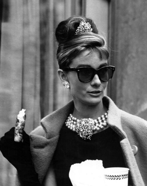 Woman from Breakfast At Tiffanys wearing sunglasses and pearls holding a pastry and coffee cup