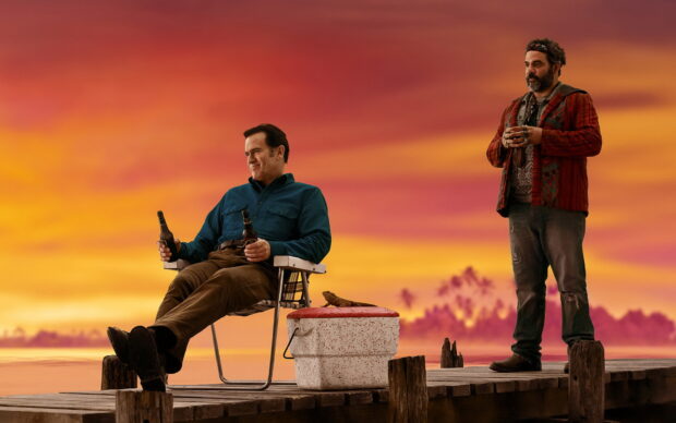 Bruce Campbell is sitting on a chair with a drink while another man stands beside him at sunset, HD Desktop Wallpaper