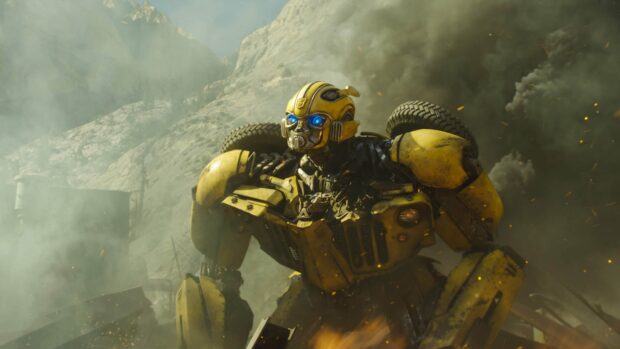 Bumblebee robot with glowing blue eyes stands amid smoke and debris in a rugged mountainous area