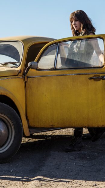 A woman stands beside a yellow vintage car under clear blue sky in this Bumblebee wallpaper